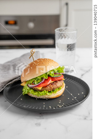 Beef burger with tomato, cheese, and lettuce on a plate on a marble table with glass of water Beef burger with tomato, cheese, and lettuce on a plate on a marble table with glass of water 139040715