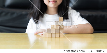 Wooden blocks and cubes form rising step shapes on a table in front of a child, combining gradual learning progress with playful problem solving and early thinking development. Wooden blocks and cubes form rising step shapes on a table in front of a child, combining gradual learning progress with playful problem solving and early thinking development. 139040897