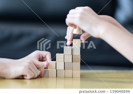 Wooden blocks and cubes form rising step shapes on a table in front of a child, combining gradual learning progress with playful problem solving and early thinking development. Wooden blocks and cubes form rising step shapes on a table in front of a child, combining gradual learning progress with playful problem solving and early thinking development. 139040899