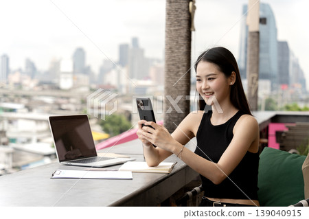 Asian adult woman uses smartphone rooftop cafe city view digital nomad online connection happy remote worker modern lifestyle global communication laptop use 139040915