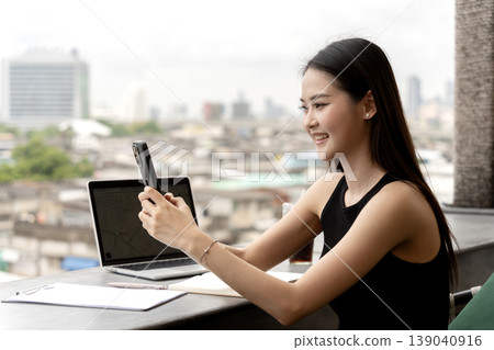 Young Asian professional woman uses smartphone video call smiling remote working cityscape cafe balcony outdoor business meeting connection 139040916