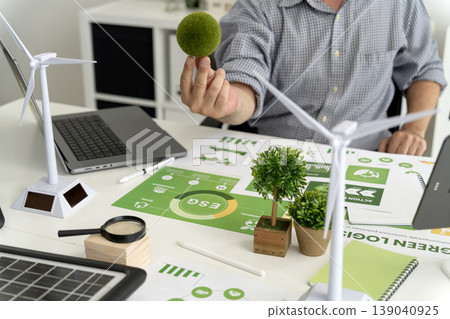 Adult male hand holding green moss ball on office desk with ESG reports wind turbines solar panel promoting sustainable business environmental strategy Adult male hand holding green moss ball on office desk with ESG reports wind turbines solar panel promoting sustainable business environmental strategy 139040925