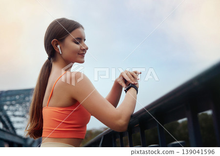 Portrait of beautiful sporty woman in sportswear using smartwatch and wireless earbud while resting Portrait of beautiful sporty woman in sportswear using smartwatch and wireless earbud while resting 139041596