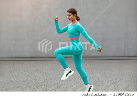 Young woman with fit body jumping and running against grey background. Female model in sportswear exercising outdoors Young woman with fit body jumping and running against grey background. Female model in sportswear exercising outdoors 139041599