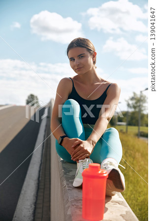 Fit woman in athletic wear sitting on a ledge beside a road, with a water bottle nearby, enjoying a moment of relaxation after an intense workout session outdoors. 139041647