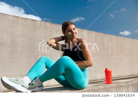 Fit woman in activewear performs a workout routine outdoors, sitting on a concrete surface with a bright orange shaker bottle beside her, showcasing dedication to fitness and healthy lifestyle. Fit woman in activewear performs a workout routine outdoors, sitting on a concrete surface with a bright orange shaker bottle beside her, showcasing dedication to fitness and healthy lifestyle. 139041654