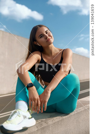 Young woman in stylish activewear poses on a ledge, showcasing a confident smile under a bright blue sky, embodying a vibrant fitness lifestyle. 139041659