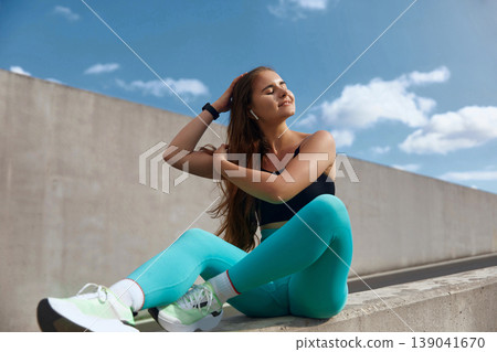 Fit woman with long hair wearing sportswear sits on a concrete ledge, enjoying the outdoors, with a bright sky and clouds in the background, embodying a healthy lifestyle and fitness concept. Fit woman with long hair wearing sportswear sits on a concrete ledge, enjoying the outdoors, with a bright sky and clouds in the background, embodying a healthy lifestyle and fitness concept. 139041670