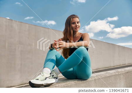 Fit young woman in athletic wear sits on a concrete ledge, showcasing a relaxed workout pose against a bright blue sky, embodying a healthy lifestyle and fitness motivation. Fit young woman in athletic wear sits on a concrete ledge, showcasing a relaxed workout pose against a bright blue sky, embodying a healthy lifestyle and fitness motivation. 139041671