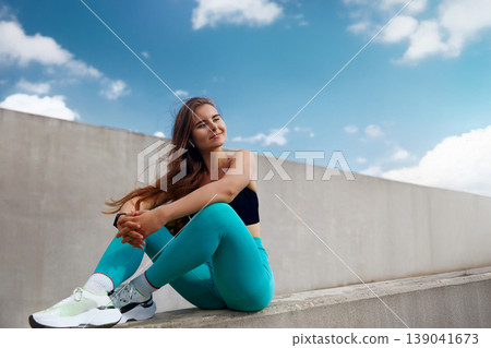 Fit young woman in athletic wear sits confidently on a ledge, showcasing vibrant turquoise leggings and stylish sneakers, embodying a sporty lifestyle and fitness motivation. Fit young woman in athletic wear sits confidently on a ledge, showcasing vibrant turquoise leggings and stylish sneakers, embodying a sporty lifestyle and fitness motivation. 139041673