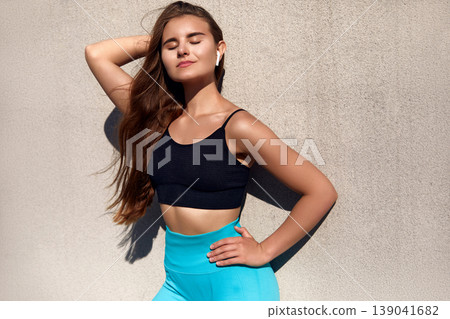 Fit young woman with long hair poses confidently in stylish activewear against a textured wall, showcasing strength and vitality in a vibrant workout environment. 139041682