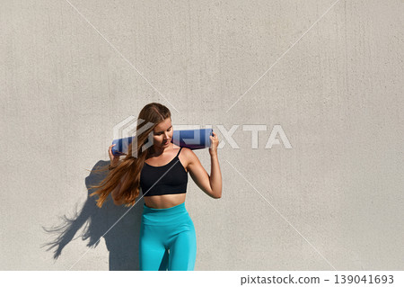 A young woman with long hair holds a purple yoga mat, wearing a black tank top and light blue leggings. She appears relaxed and confident as she approaches an exercise space A young woman with long hair holds a purple yoga mat, wearing a black tank top and light blue leggings. She appears relaxed and confident as she approaches an exercise space 139041693