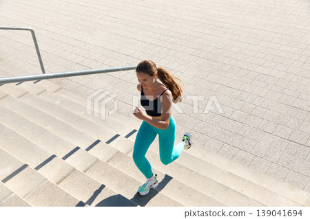 Athletic woman running up to the stone stairs, using wireless headphones in City with fitness tracker. Athletic woman running up to the stone stairs, using wireless headphones in City with fitness tracker. 139041694