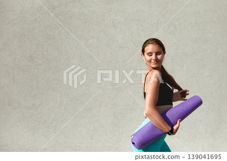 Woman Carrying a Yoga Mat Walks Confidently Against a Minimalistic Background During a Sunny Day Woman Carrying a Yoga Mat Walks Confidently Against a Minimalistic Background During a Sunny Day 139041695
