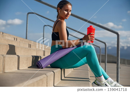 Fit woman sitting on outdoor steps, holding a red shaker bottle, with a yoga mat beside her, showcasing a moment of relaxation after a workout in a sporty environment. 139041718