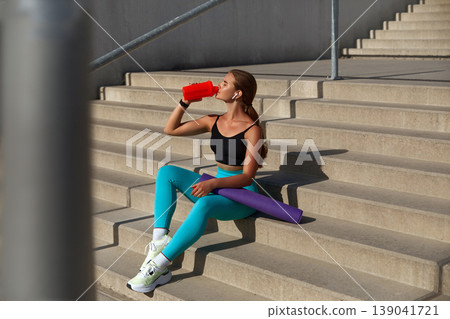 Fit young woman in athletic wear sits on concrete steps, sipping from a red water bottle, with a yoga mat beside her, showcasing a refreshing workout break in an urban environment. Fit young woman in athletic wear sits on concrete steps, sipping from a red water bottle, with a yoga mat beside her, showcasing a refreshing workout break in an urban environment. 139041721