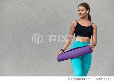 A young woman with long hair dressed in a black sports top and turquoise leggings stands against a textured concrete wall, holding a yoga mat and representing a healthy fitness lifestyle. 139041737