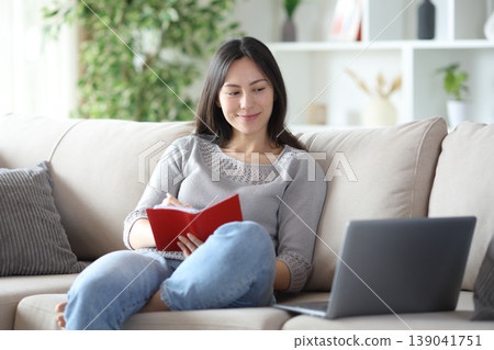 Asian woman writing in agenda checking computer at home Asian woman writing in agenda checking computer at home 139041751