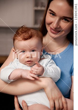 Mother holding baby in her arms while smiling at the camera in a cozy indoor setting during daytime 139041941