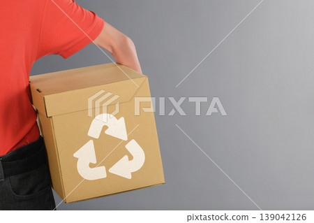 A person holding a brown cardboard box featuring a white recycling logo against a neutral gray background, representing environmental sustainability and waste reduction 139042126
