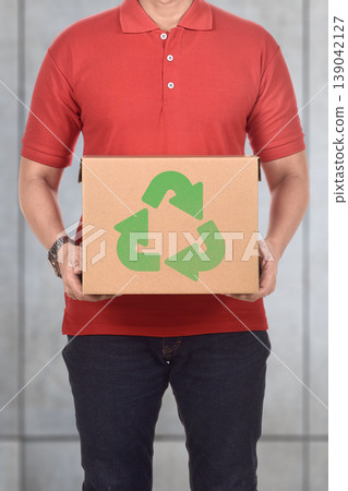 A man in a red shirt holds a cardboard box featuring a green recycling logo, representing environmental sustainability and eco-friendly packaging practices 139042127