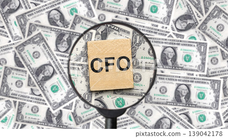 A wooden block with CFO text under a magnifying glass on a background of US dollar bills. Represents chief financial officer, accounting, auditing, and corporate finance management 139042178
