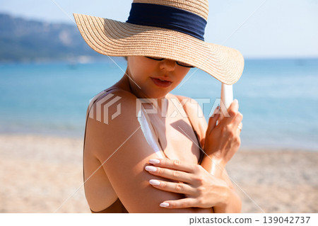 Pretty Woman Applying Sunscreen On Shoulder With Hat At Beach. Beauty Skincare Concept. Female Using Sun Cream For Healthy Summer Skin 139042737