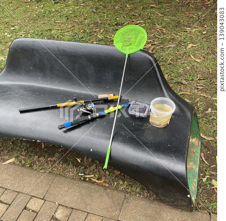 Fishing gear and container set on a bench at a park during the day with grass nearby 139043083