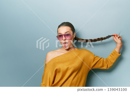 Beautiful woman holding braid posing playfully in yellow sweater and sunglasses on blue background expressing fun lifestyle emotion and modern beauty portrait energy Beautiful woman holding braid posing playfully in yellow sweater and sunglasses on blue background expressing fun lifestyle emotion and modern beauty portrait energy 139043100