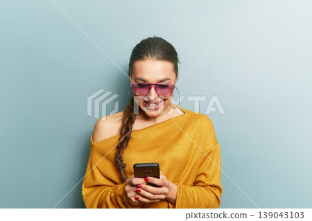 Young woman smiling and using smartphone in yellow sweater and sunglasses on blue background representing digital lifestyle communication and modern beauty portrait Young woman smiling and using smartphone in yellow sweater and sunglasses on blue background representing digital lifestyle communication and modern beauty portrait 139043103