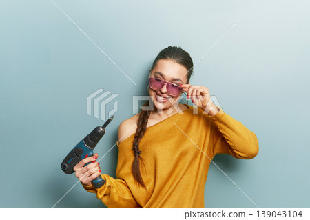 Beauty woman holding drill tool playfully in yellow sweater and sunglasses on blue background mixing DIY concept with fashion lifestyle and creative portrait Beauty woman holding drill tool playfully in yellow sweater and sunglasses on blue background mixing DIY concept with fashion lifestyle and creative portrait 139043104