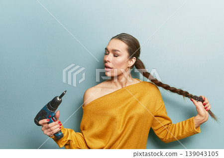 Female model holding drill and braid looking sideways in yellow sweater on blue background expressing creative DIY concept and modern lifestyle beauty portrait 139043105