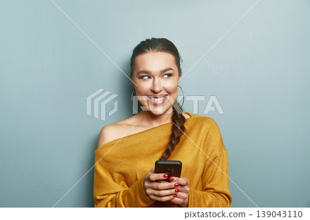 Beauty woman smiling gently holding smartphone in yellow sweater on blue background expressing calm lifestyle digital connection and soft beauty portrait 139043110