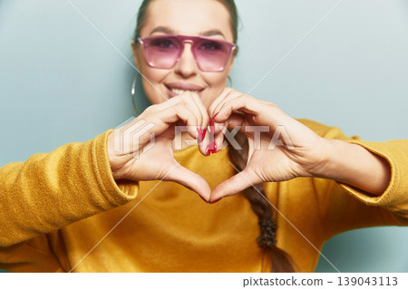 Attractive woman close up making heart hands gesture wearing sunglasses and yellow sweater on blue background expressing love concept and confident beauty portrait Attractive woman close up making heart hands gesture wearing sunglasses and yellow sweater on blue background expressing love concept and confident beauty portrait 139043113