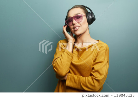Woman listens to music with headphones while smiling in a studio against a plain blue backdrop during daytime Woman listens to music with headphones while smiling in a studio against a plain blue backdrop during daytime 139043123