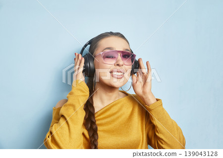 Woman enjoys music while wearing headphones and sunglasses in front of a blue wall with a bright smile during daytime Woman enjoys music while wearing headphones and sunglasses in front of a blue wall with a bright smile during daytime 139043128