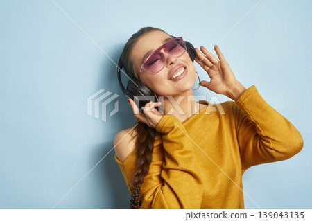 Young woman enjoys music while wearing headphones and sunglasses against a blue background during a casual setting 139043135