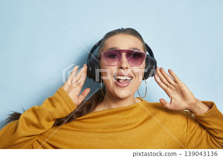 Excited young woman smiles while listening to music on headphones against a blue wall during a sunny day 139043136