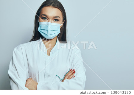 Health worker wearing a mask stands with arms crossed in a clinical setting during the day Health worker wearing a mask stands with arms crossed in a clinical setting during the day 139043150
