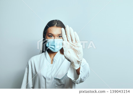 Medical professional wearing gloves and a mask gestures to stop while standing against a plain background Medical professional wearing gloves and a mask gestures to stop while standing against a plain background 139043154