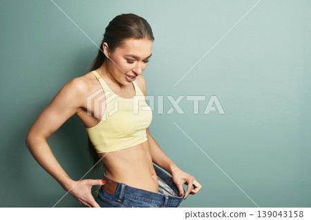 Woman shows her success in weight loss by fitting into larger jeans while standing against a simple background in a room Woman shows her success in weight loss by fitting into larger jeans while standing against a simple background in a room 139043158