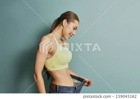 Woman smiles while holding oversized pants in front of green wall during indoor activity showcasing weight loss achievement Woman smiles while holding oversized pants in front of green wall during indoor activity showcasing weight loss achievement 139043162