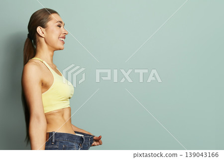 Woman smiles while holding large jeans that no longer fit in a light green room during a fitness journey 139043166