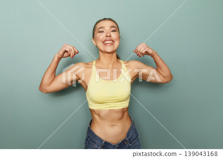 Fit woman shows her muscles in front of a green wall during a workout session at a gym while smiling and expressing confidence in her strength and fitness journey Fit woman shows her muscles in front of a green wall during a workout session at a gym while smiling and expressing confidence in her strength and fitness journey 139043168