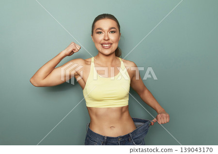 Woman shows weight loss success standing against green wall wearing yellow top and blue jeans after fitness journey and healthy changes in lifestyle Woman shows weight loss success standing against green wall wearing yellow top and blue jeans after fitness journey and healthy changes in lifestyle 139043170