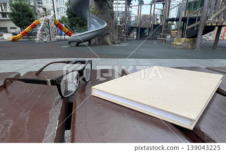 Glasses and a notebook are left on a wooden bench at a playground with slides Glasses and a notebook are left on a wooden bench at a playground with slides 139043225