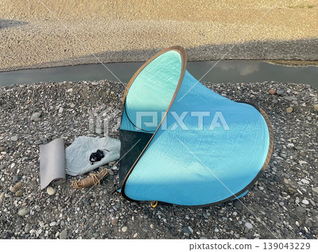 Tent is set up near a riverbank on a gravel area during daylight with some items laid out nearby Tent is set up near a riverbank on a gravel area during daylight with some items laid out nearby 139043229