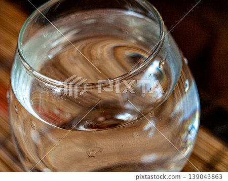 Glass of water with refraction of a wooden table texture. Macro shot featuring optical distortion of wood grain and rhythmic patterns through a transparent liquid cylinder in soft light Glass of water with refraction of a wooden table texture. Macro shot featuring optical distortion of wood grain and rhythmic patterns through a transparent liquid cylinder in soft light 139043863