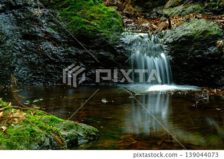 A small waterfall flows through a silent forest, surrounded by moss-covered rocks and pure spring water. A small waterfall flows through a silent forest, surrounded by moss-covered rocks and pure spring water. 139044073