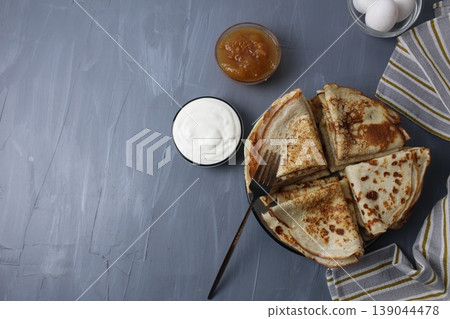 Crepe pancakes folded into triangles lying on a plate with sour cream and jam in a gravy boat on a gray background with space for text 139044478